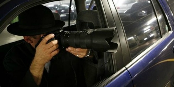 Person wearing a black hat holding a large camera with a telephoto lens, taking a photo through the open window of a blue car.