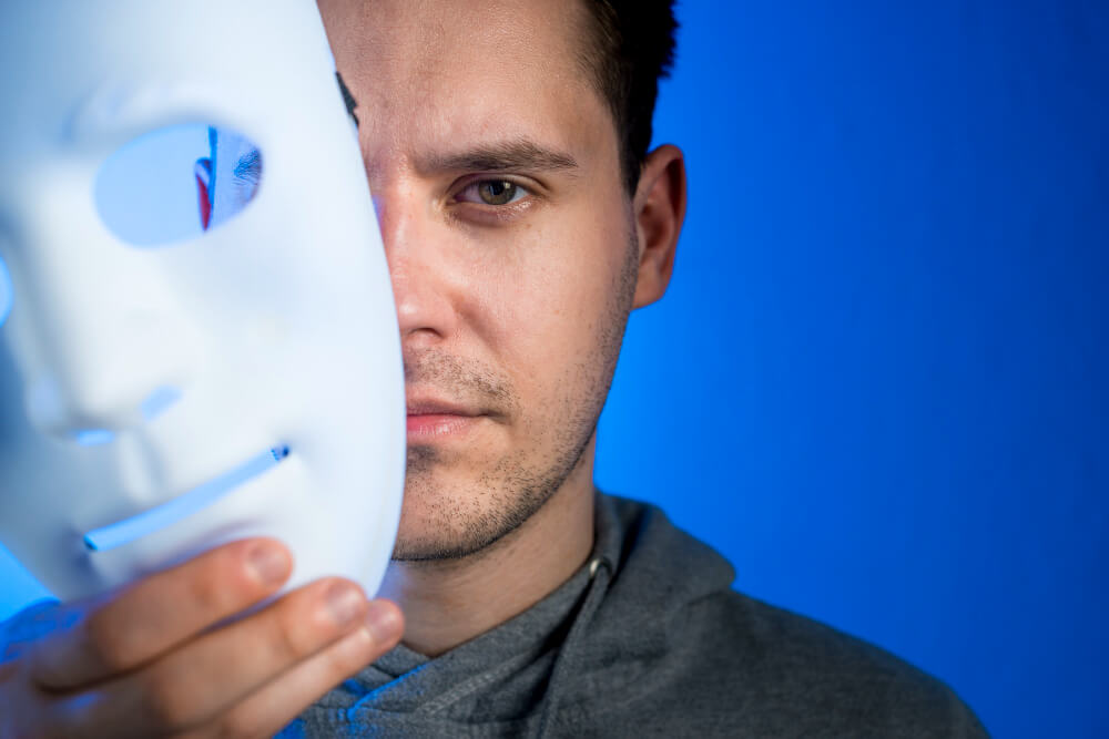 Man holding a white theatrical mask partially covering his face against a blue background.