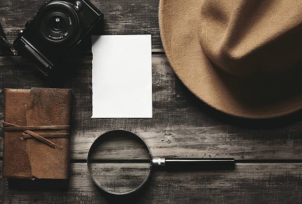 Vintage camera, blank white paper, brown leather notebook, magnifying glass, and tan felt hat on a rustic wooden surface.