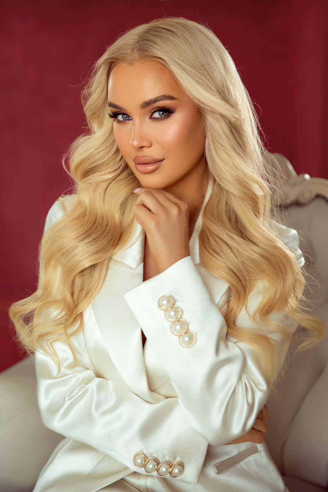 Kobieta z długimi blond falowanymi włosami w białym żakiecie z perłowymi guzikami, na tle bordowym.