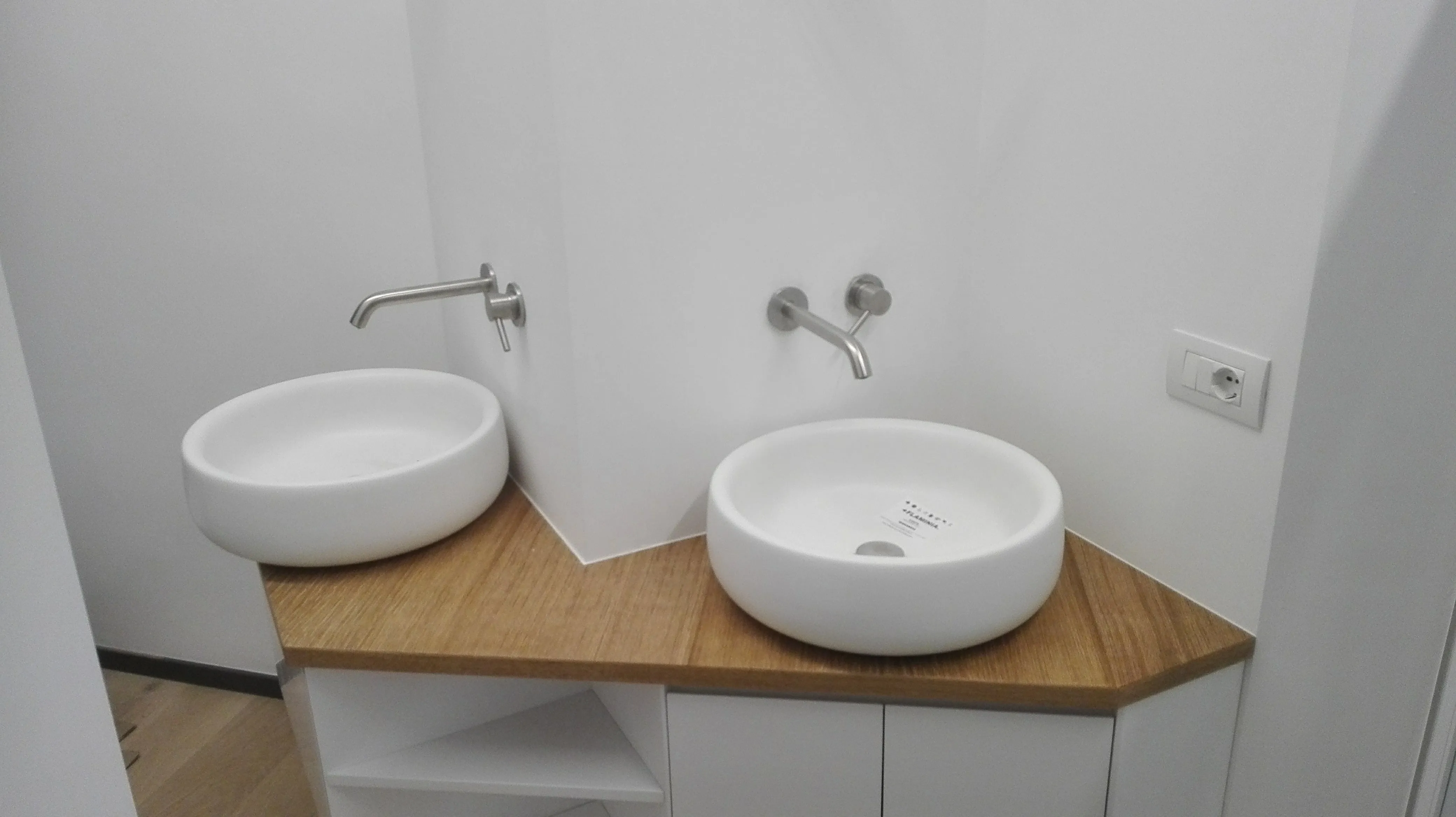 Due lavandini bianchi rotondi su un piano angolare in legno con rubinetti a muro in bagno.