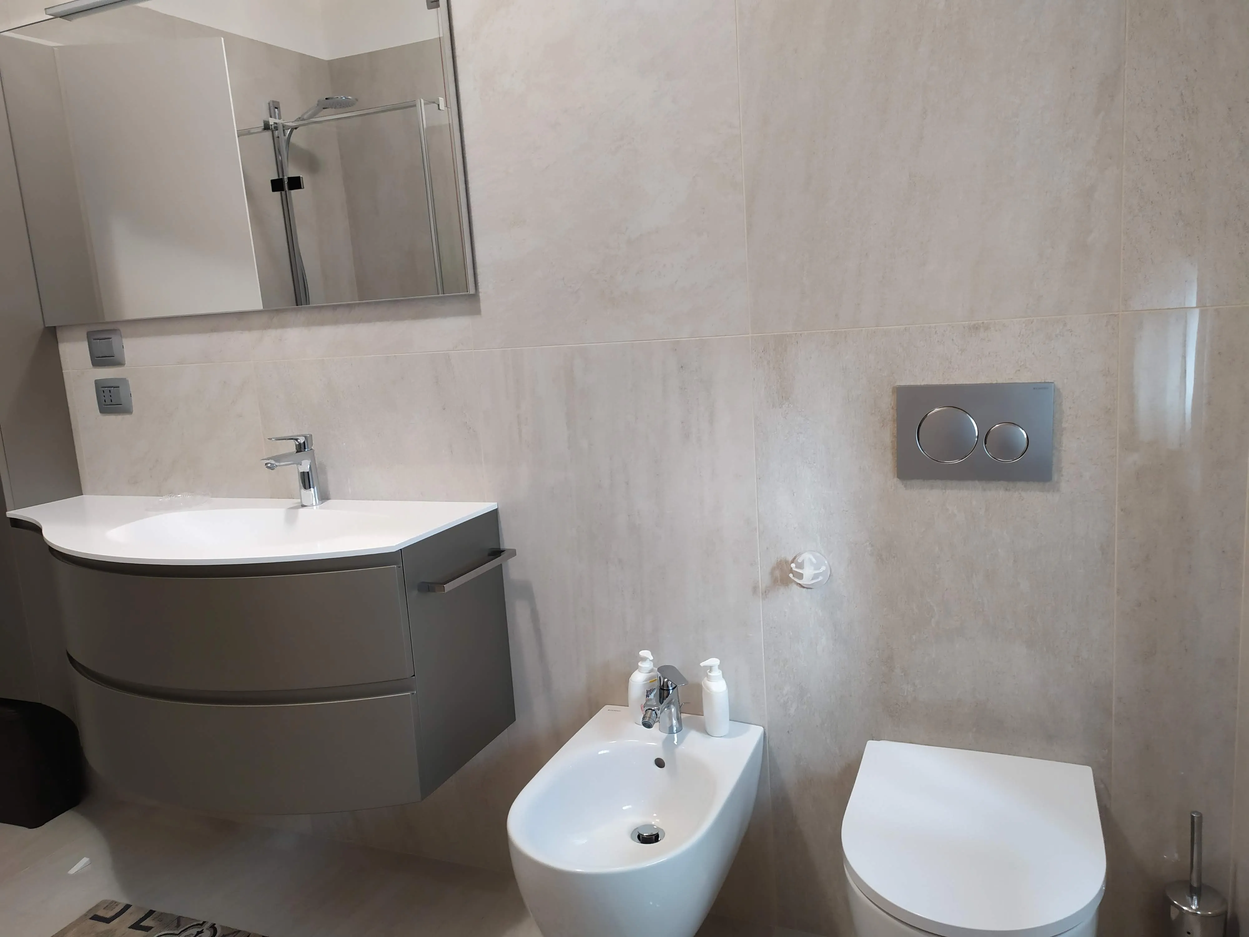 Bagno moderno con lavabo sospeso, bidet con sapone e dispenser, e WC con pulsanti di scarico a parete.