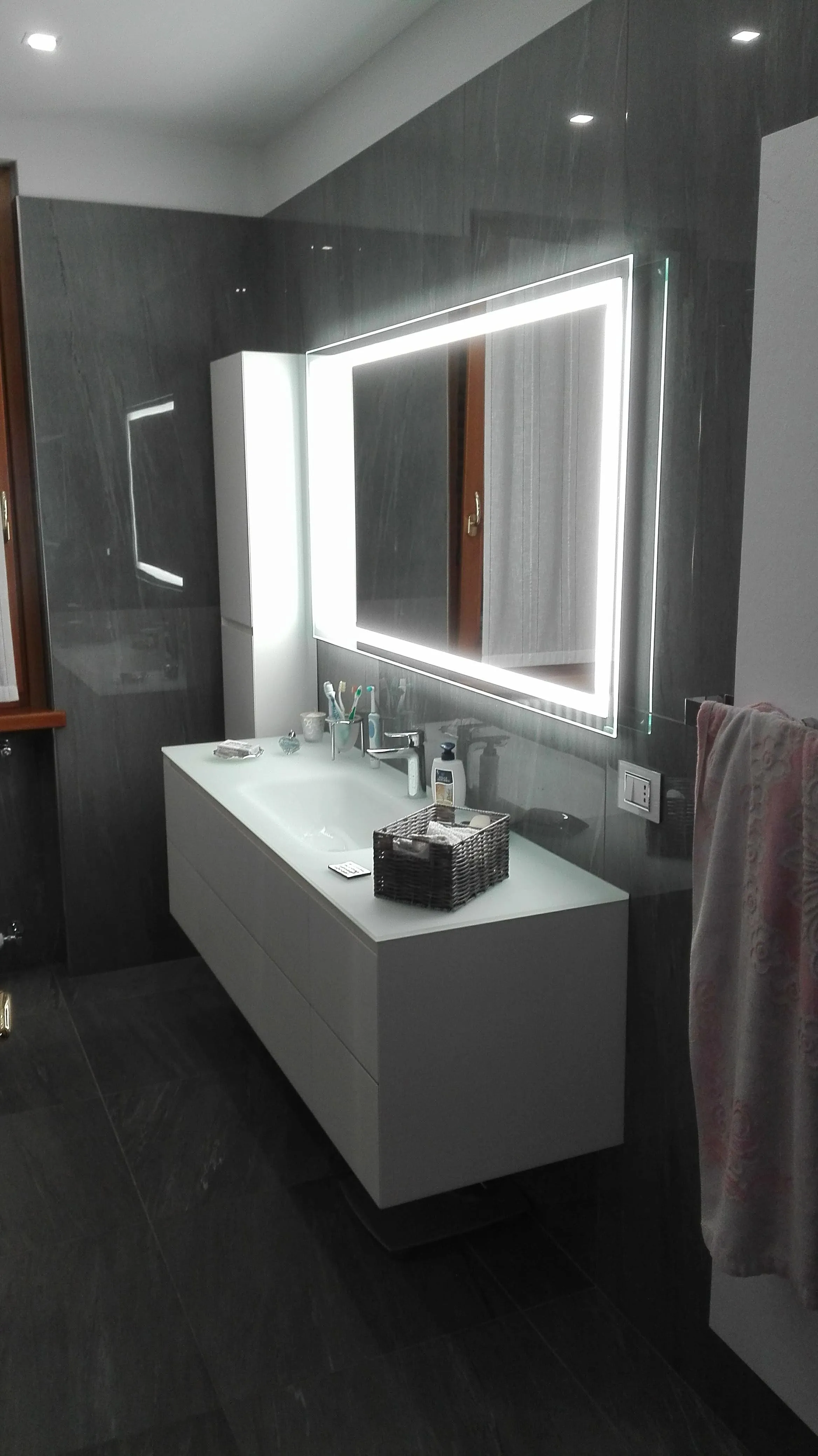 Bagno moderno con lavabo bianco, specchio illuminato, mobile contenitore e pareti grigie lucide.