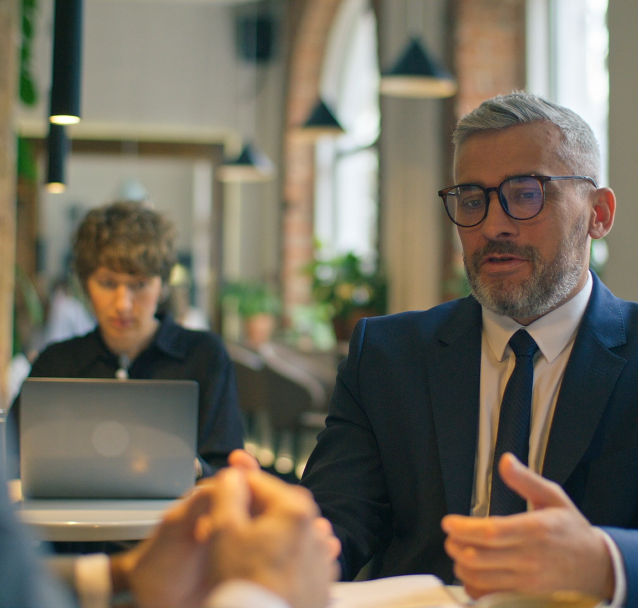 Man in suit and glasses talking in a professional setting with blurred person typing on a laptop in the background.