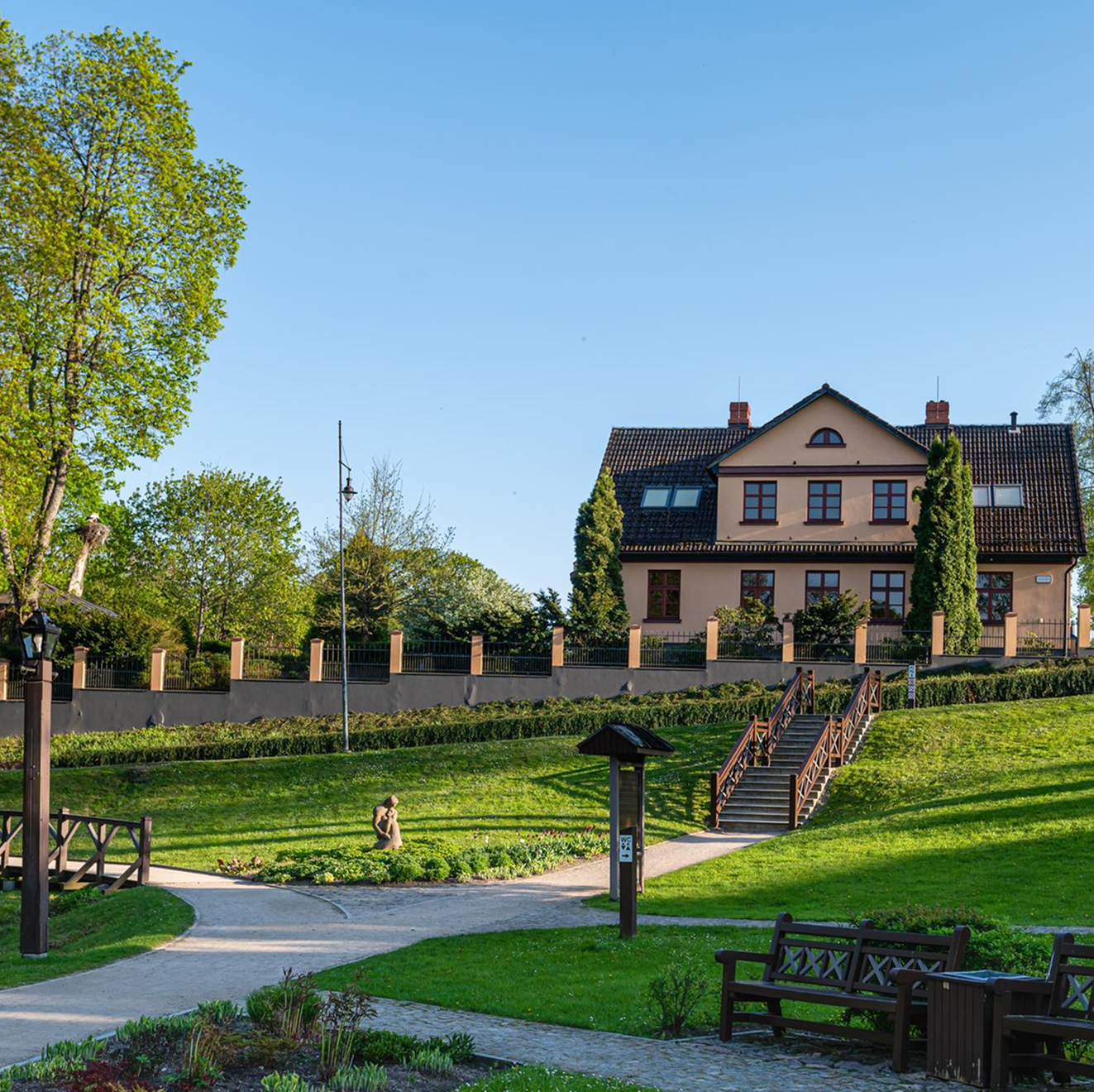 A large house with a tiled roof surrounded by green lawns, trees, and a pathway with benches in a park-like setting under a clear blue sky.