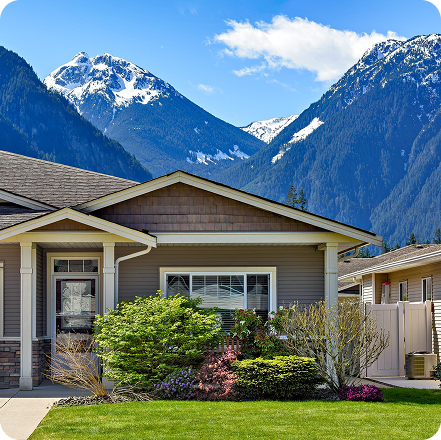 Single-story beige house with landscaped front yard and snow-capped mountains in the background under a blue sky.