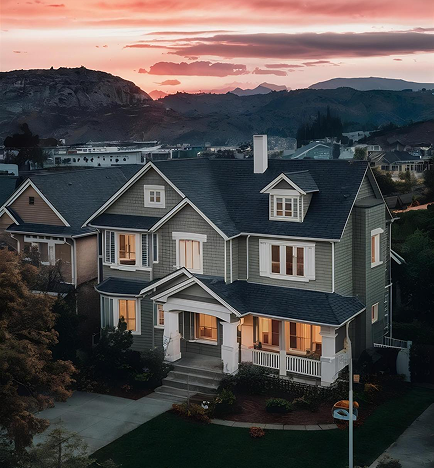 Large two-story gray house with lit windows at dusk, surrounded by greenery and hills in the background.