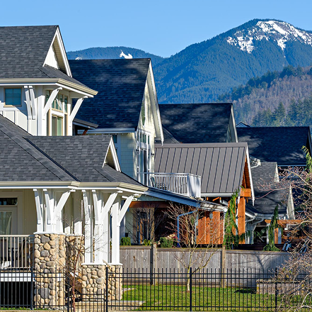 Row of modern houses with stone pillars and black shingle roofs in front of snow-capped mountains under a clear blue sky.