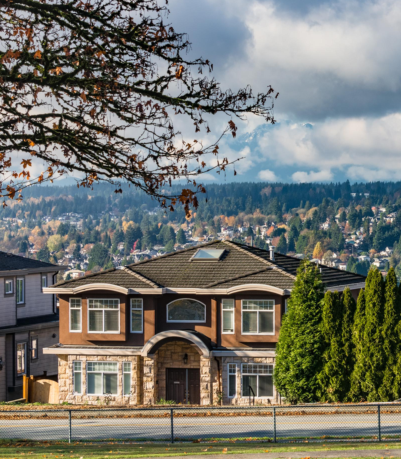 Two-story house with stone and brown siding, large windows, surrounded by tall evergreen trees and distant forest-covered hills under cloudy sky.