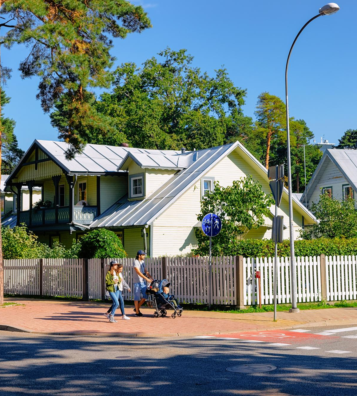Family walking on a sidewalk with a stroller near white wooden houses and green trees on a sunny day.