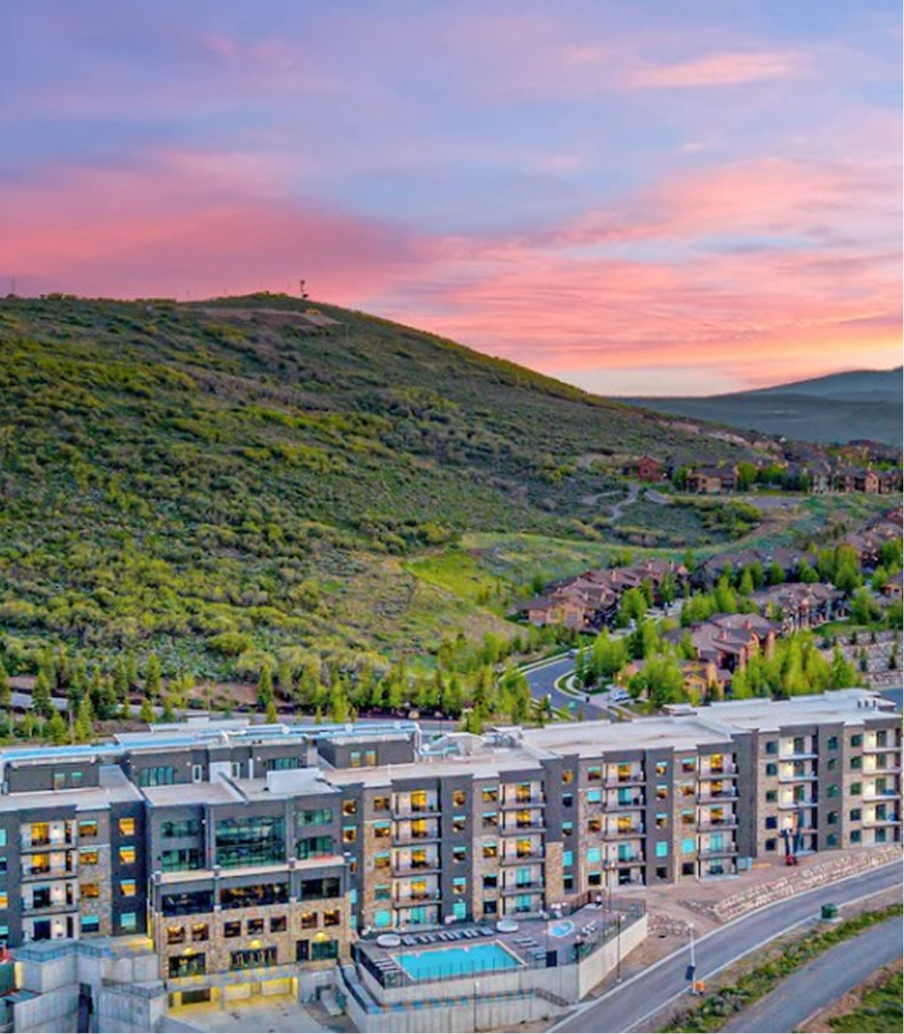 A modern multi-story apartment complex with a swimming pool at sunset beside a green hillside and residential houses.