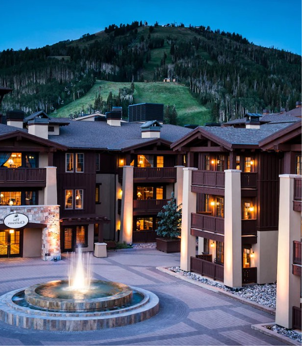 Courtyard of a mountain lodge with a lit fountain, balconies, and forested slopes in the background at dusk.
