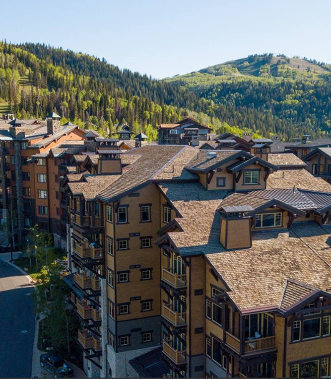 A cluster of brown mountain lodges with balconies under clear blue sky, surrounded by forested hills.