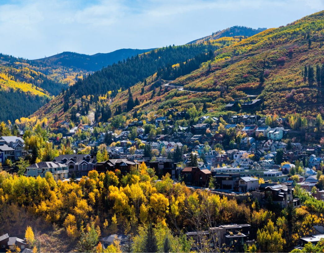 Mountain town surrounded by autumn trees with yellow and green foliage under a clear blue sky.