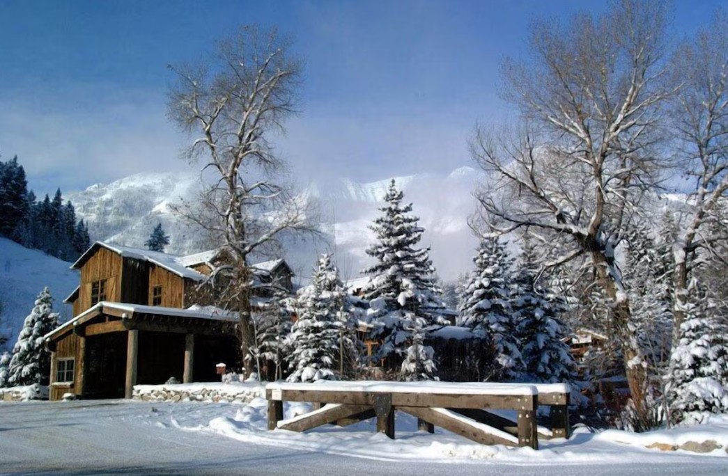 Snow-covered wooden cabin surrounded by snow-laden trees and a wooden fence with snowy mountains in the background under a clear blue sky.
