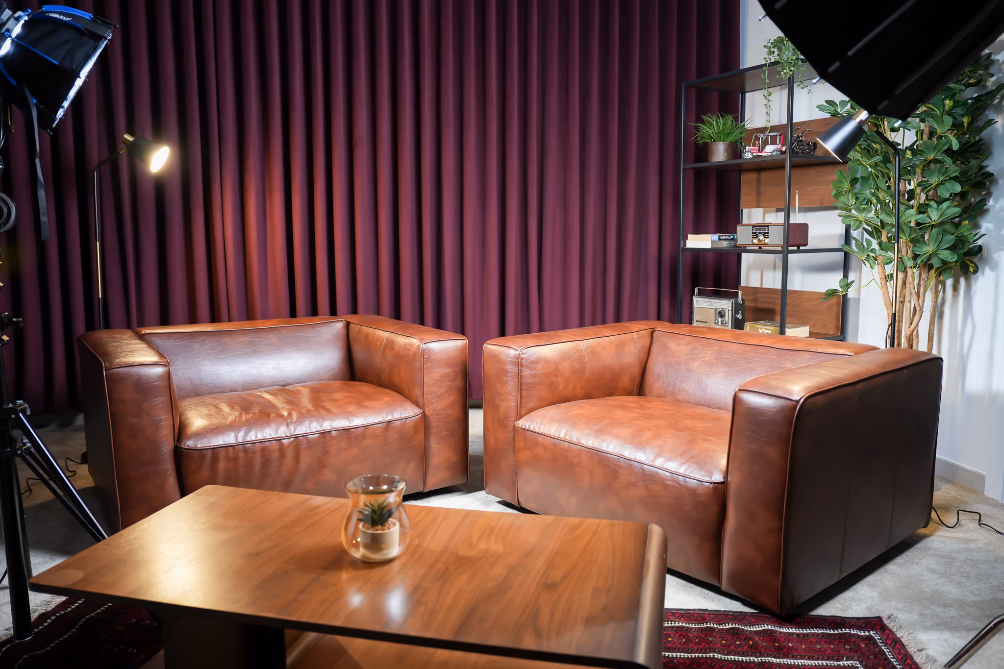2 brown leather chairs and a brown table made in retro style with a maroon curtain and a shelf in the background.