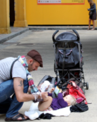 Eltern spielen draußen mit ihren Kindern – ein Mann hält ein Kind kopfüber, während eine Frau ein Baby in einer Decke trägt, in einer natürlichen Umgebung.