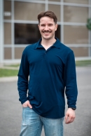 Homme souriant portant un polo bleu marine à manches longues et un jean clair debout devant un bâtiment moderne avec de grandes fenêtres.
