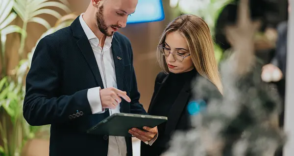 Un uomo con giacca scura mostra qualcosa su un tablet a una donna con occhiali e maglione nero, entrambi sono concentrati sullo schermo.