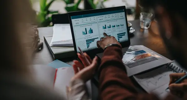 Due persone che lavorano su un tablet con grafici e statistiche, una indica lo schermo mentre l'altra prende appunti su un taccuino accanto a documenti e libri aperti su un tavolo.