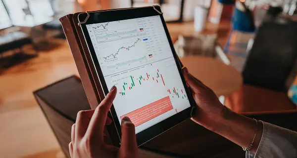 Una persona tiene in mano un tablet che mostra grafici finanziari con linee di tendenza e indicatori, mentre si trova in un ambiente interno luminoso con mobili sullo sfondo.