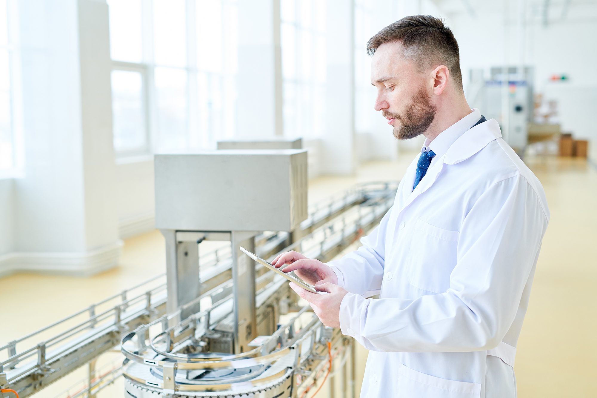 Profile view of confident bearded machine operator wearing white coat using digital tablet while wrapped up in work at modern production department of dairy plant, portrait shot