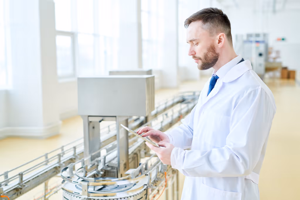 Profile view of confident bearded machine operator wearing white coat using digital tablet while wrapped up in work at modern production department of dairy plant, portrait shot