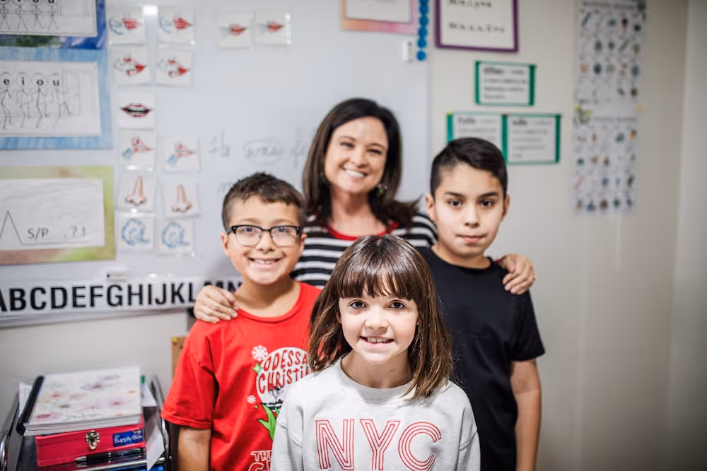 Three children smiling with a female teacher standing behind them in a classroom with educational posters on the wall.