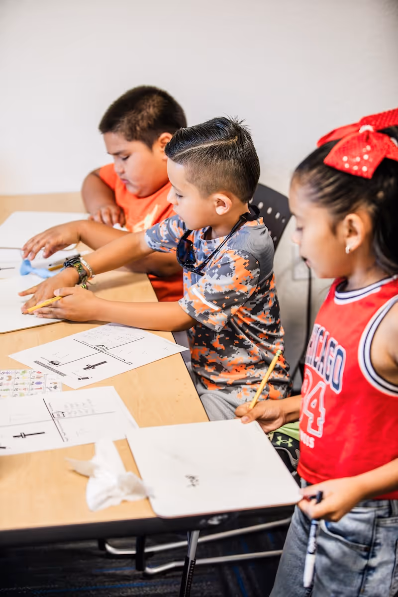 Three children working at a table with pencils and worksheets, focused on their tasks in a classroom setting.
