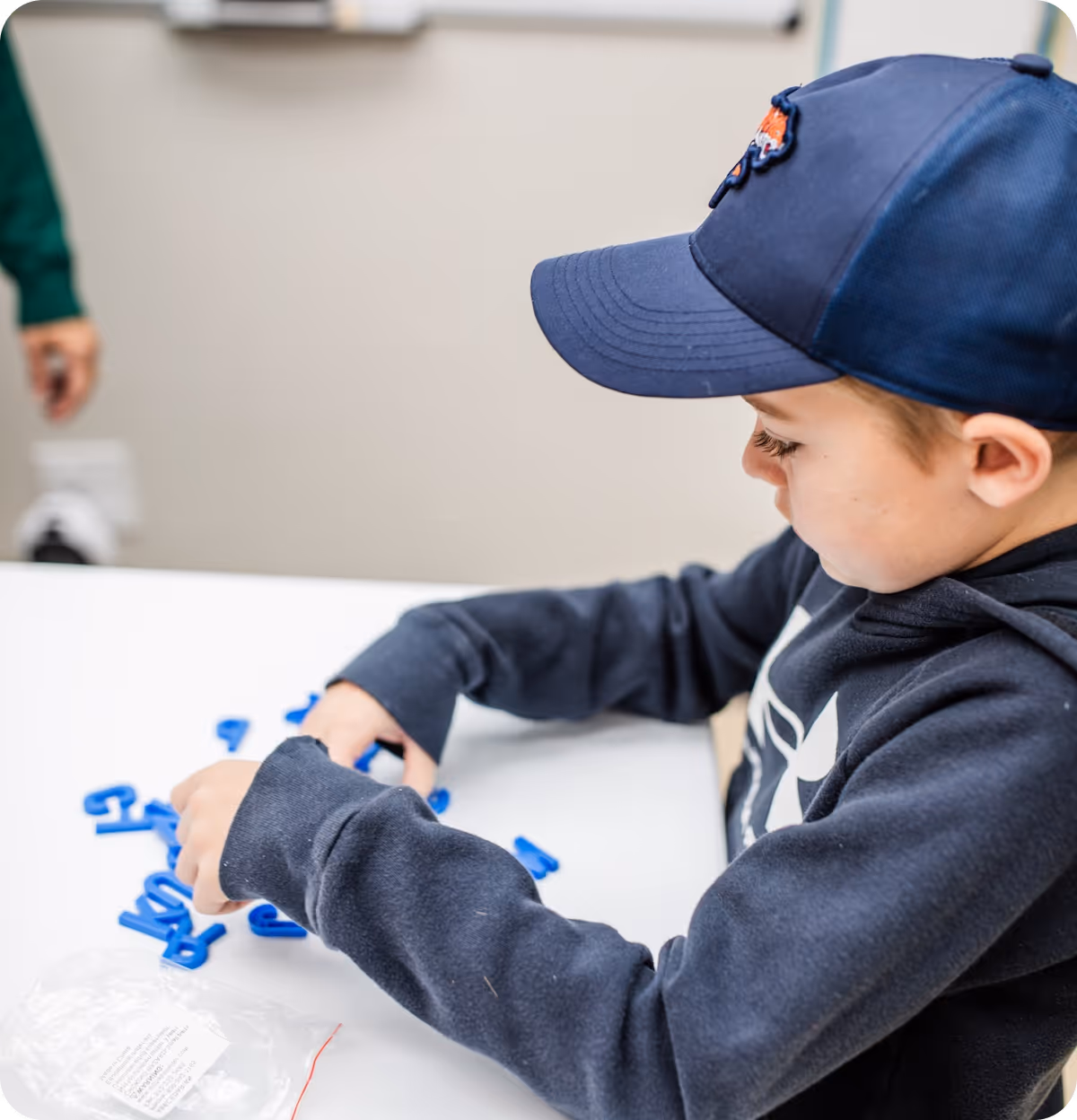 Young boy wearing a dark blue cap and hoodie arranging blue plastic letters on a white table.