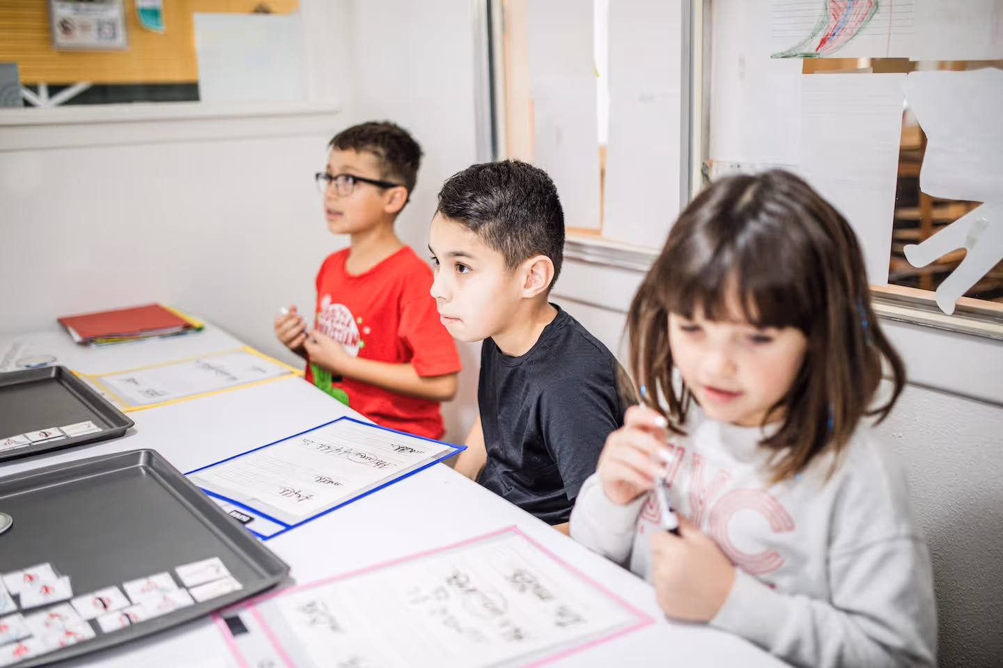 Three children sitting at a table engaged in a classroom activity with worksheets and trays in front of them.