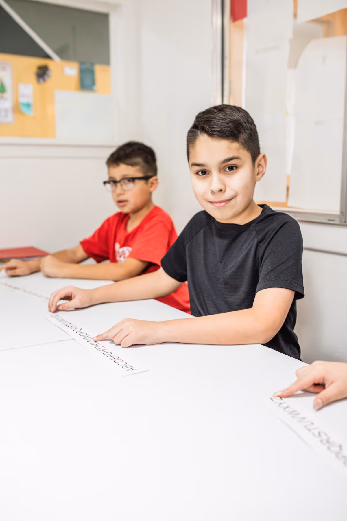 Two boys sitting at a table pointing to printed alphabet strips during a classroom activity.