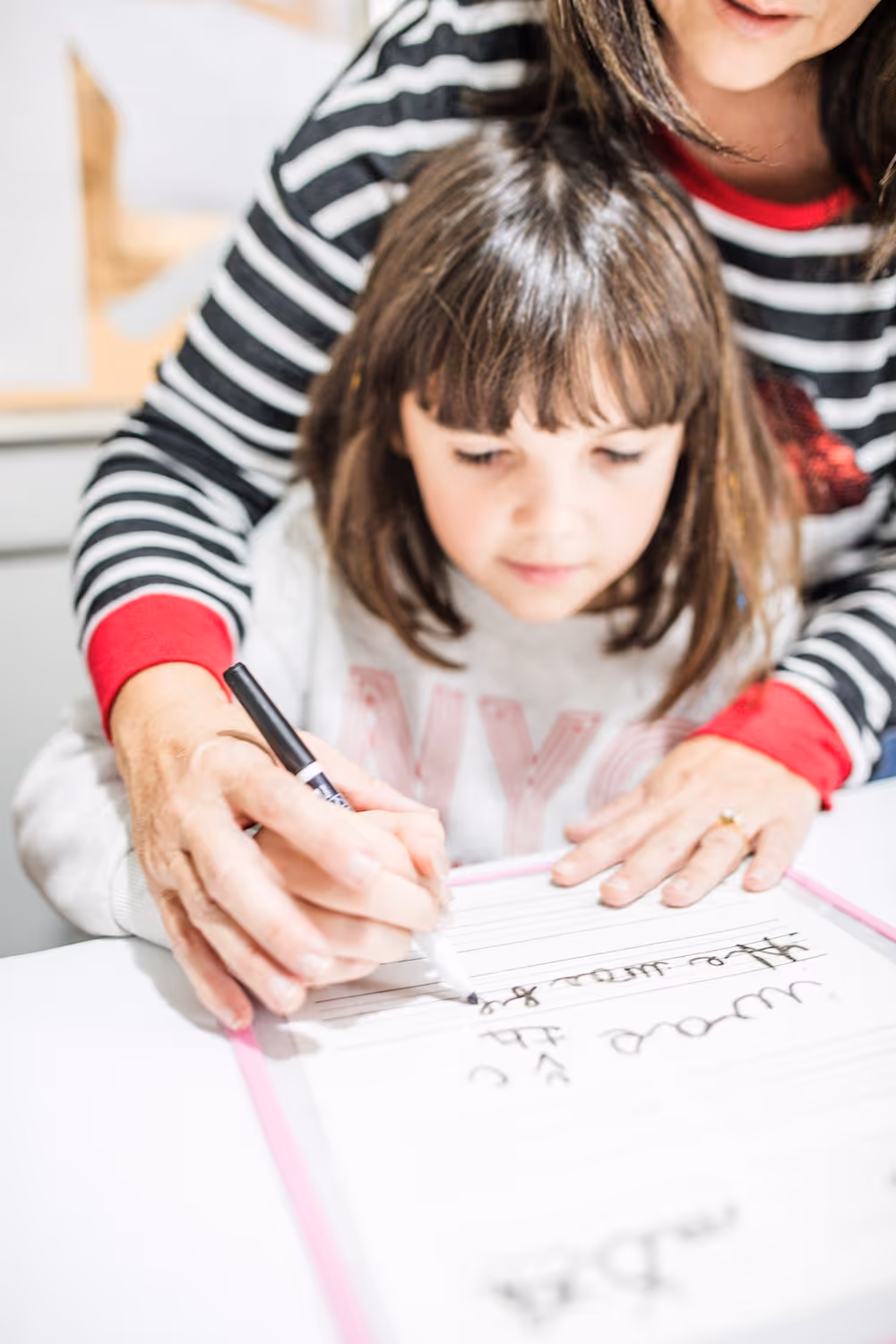 Child being guided by adult hands to write on a whiteboard with cursive letters.