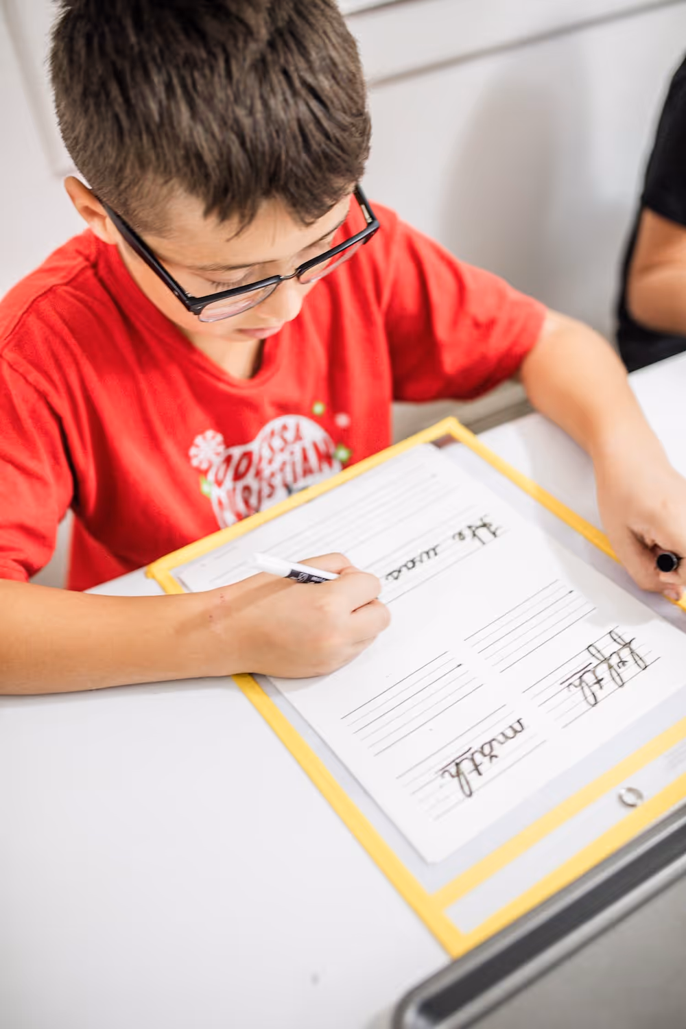Boy with glasses in red shirt practicing handwriting on lined paper at a desk.