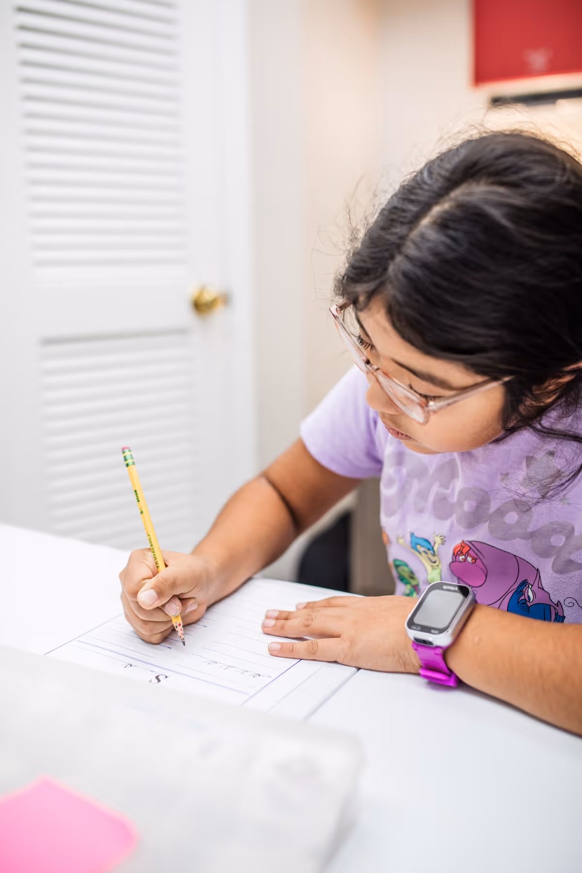 Child wearing glasses and a purple cartoon t-shirt writing with a pencil on a sheet of paper at a white desk.