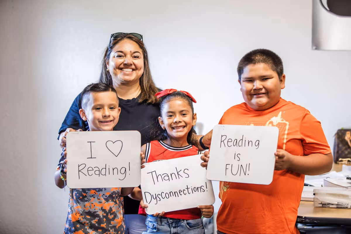 Smiling woman and three children holding signs that say 'I ♥ Reading!', 'Thanks Dysconnections!', and 'Reading is FUN!' in a bright room.