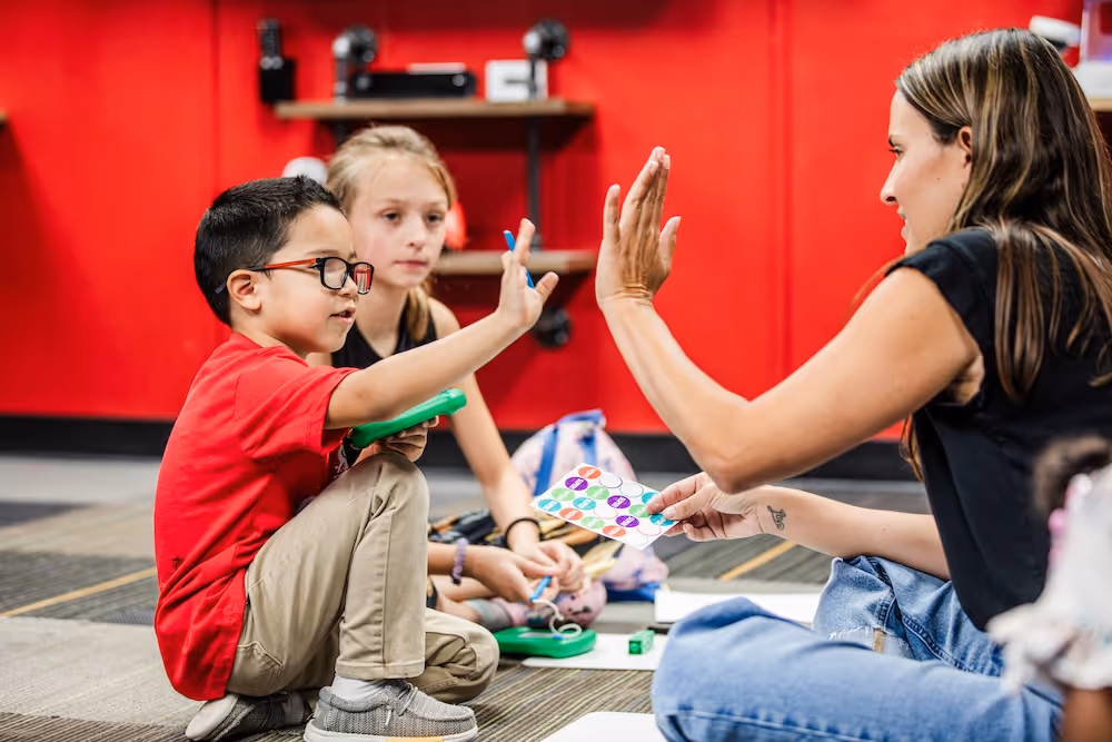 Young boy in red shirt giving a high-five to a woman while sitting on the floor in a classroom setting.