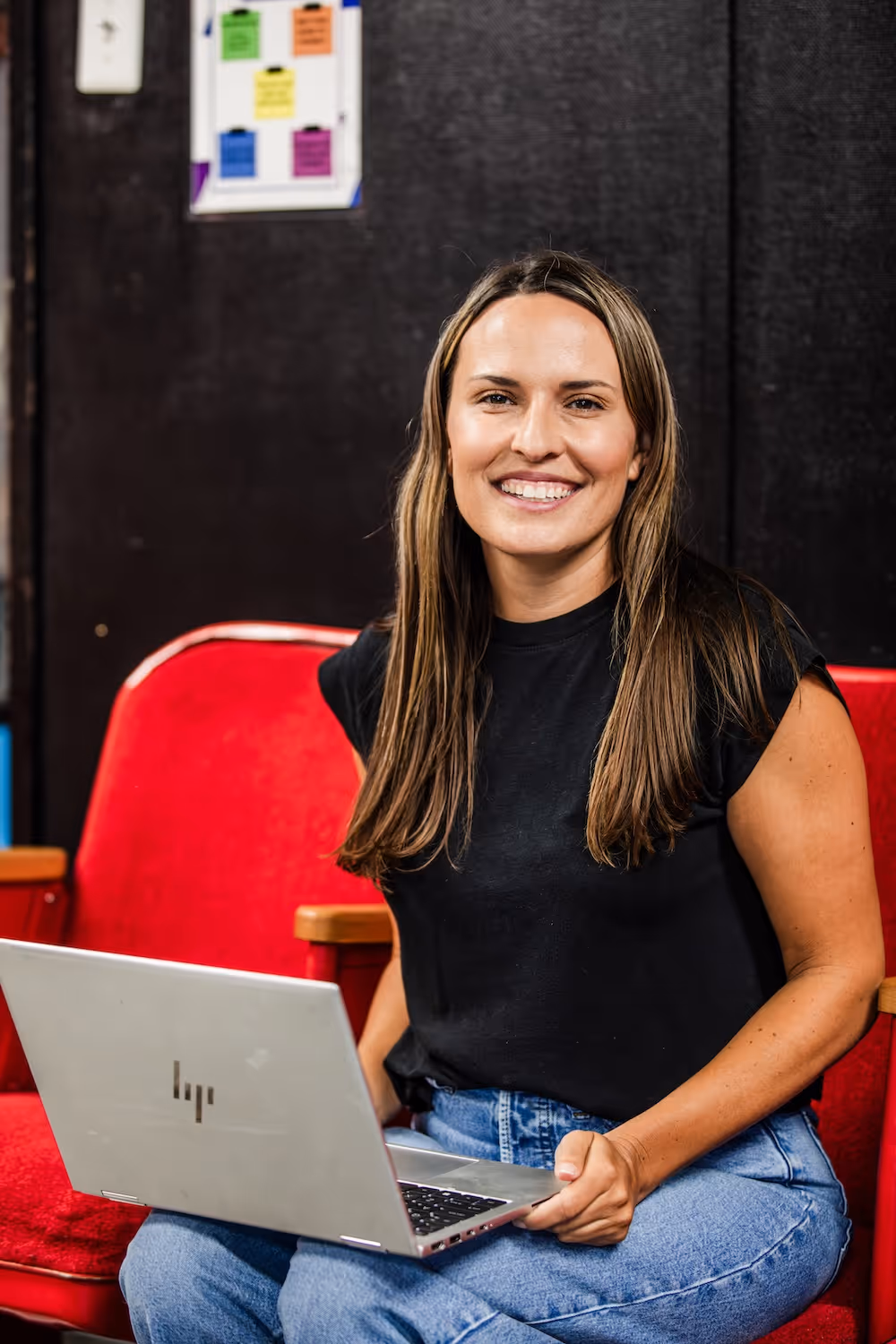 Smiling woman with long brown hair wearing a black t-shirt and blue jeans sitting on a red chair with a laptop on her lap.