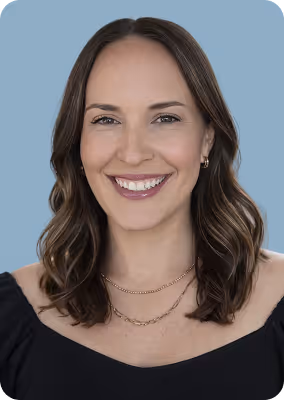 Smiling woman with shoulder-length wavy brown hair wearing a black top and layered gold necklaces against a blue background.