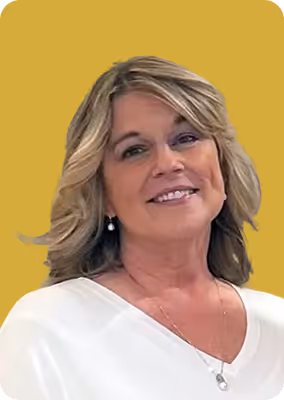 Smiling woman with blonde hair wearing pearl earrings and white top against a mustard yellow background.