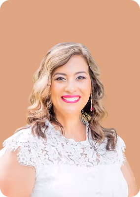 Smiling woman with wavy blonde and brown hair wearing a white lace top and dangling earrings against a beige background.