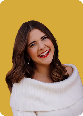 Smiling woman with long brown hair wearing red lipstick and a white off-shoulder sweater against a mustard yellow background.