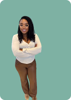 Smiling woman with glasses, wearing a white long-sleeve top and brown pants, standing with arms crossed against a green background.