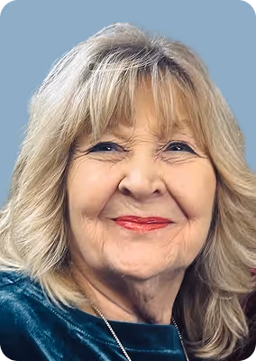 Smiling older woman with blonde hair and red lipstick wearing a green velvet top against a blue background.