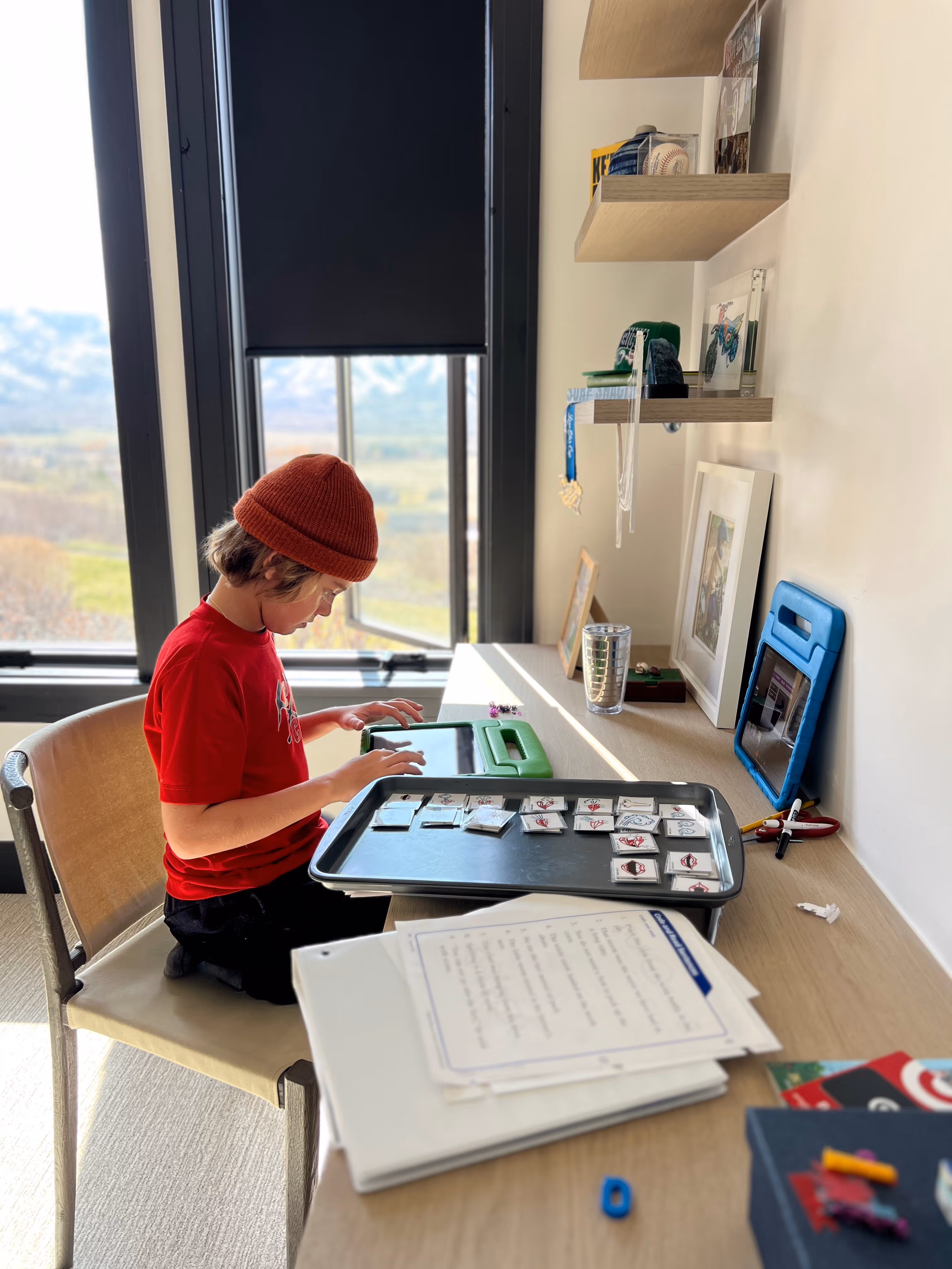 Child wearing red shirt and hat uses a tablet at a desk with papers and learning materials.