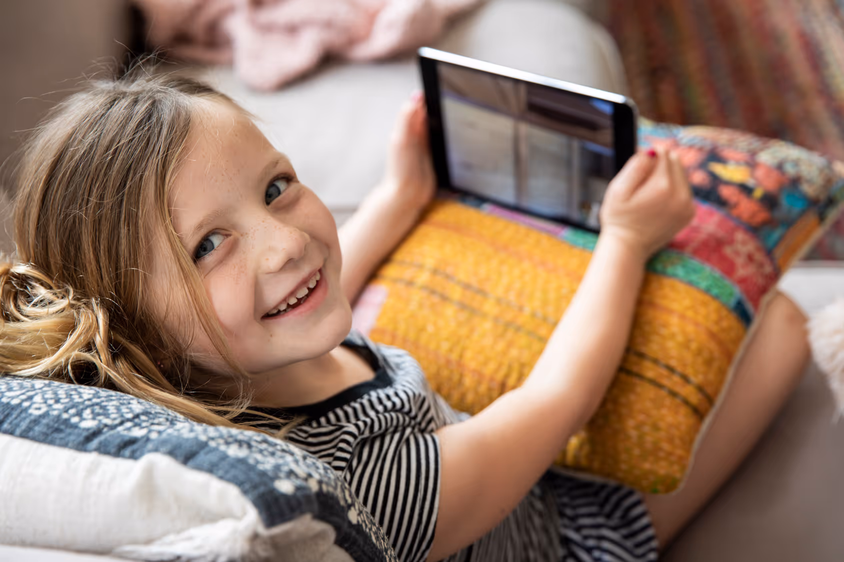 Smiling young girl lying on couch, holding a tablet and resting on a colorful pillow.