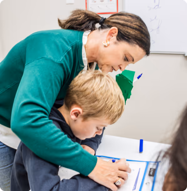 Teacher in a green sweater closely assisting a young blond boy with writing at a desk.