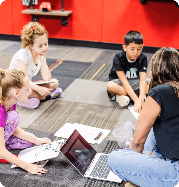 A teacher with three children sitting on a patterned carpet floor engaged in a group activity with clipboards and a laptop.