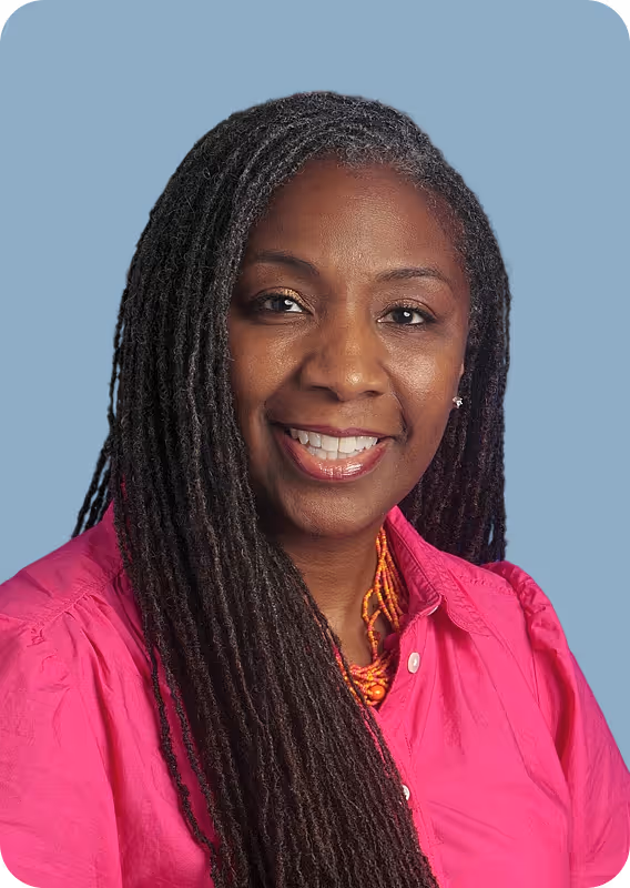 Smiling woman with long dreadlocks wearing a bright pink shirt and orange necklace against blue background.