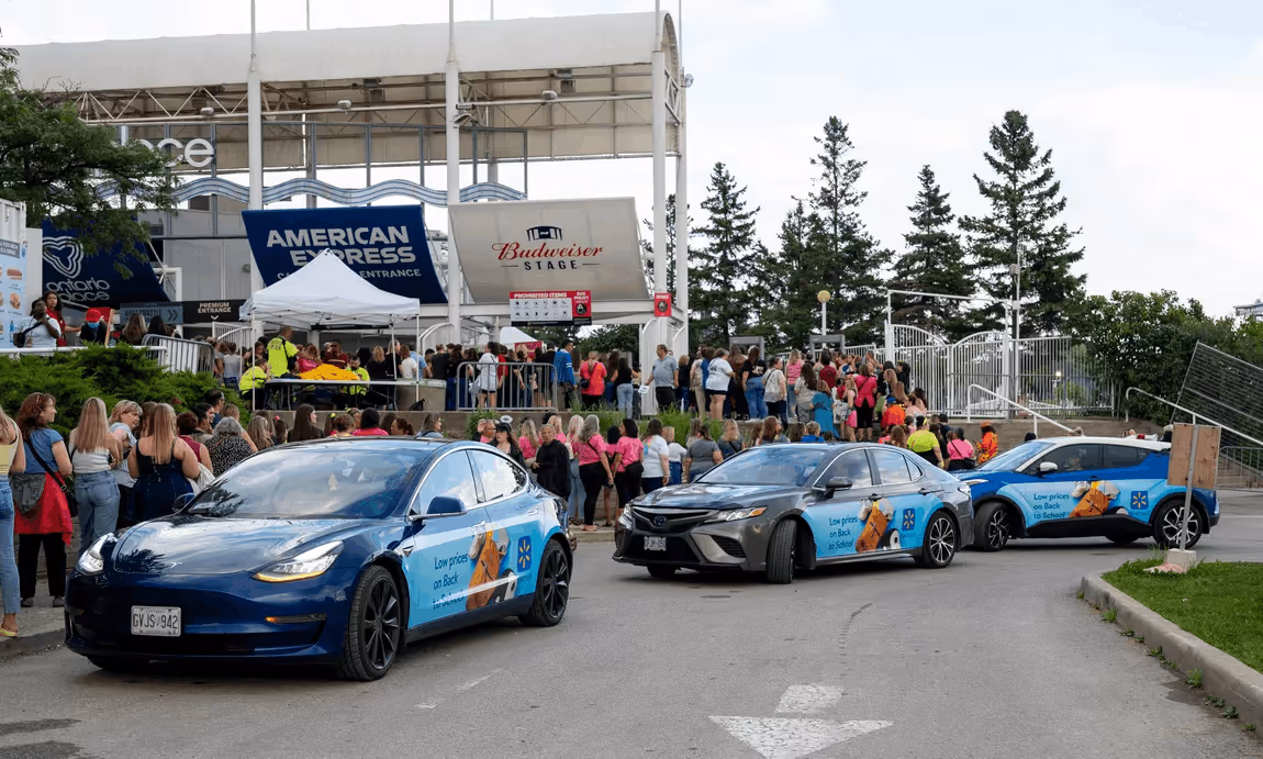 Three blue cars with Walmart advertisements parked near a crowd outside the American Express and Budweiser Stage entrance.